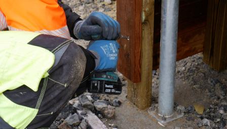 Suojavarusteisiin pukeutunut rakennustyöntekijä kiinnittää lautaan naulaa porakoneella.