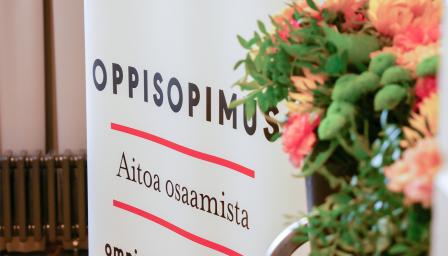 Taustalla roll-up, jossa teksti Oppisopimus, aitoa osaamista. Etualalla kukkia.
