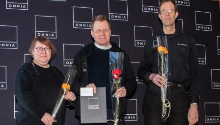 Kolme ihmistä katsoo kameraan käsissään kukat ja kunniakirja.