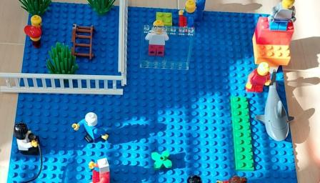 Legorakennelma, jossa on erilaisia legohahmoja. Rakennelmassa osa hahmoista on sisällä ja osa ulkona. Erilaiset tilat on kuvattu legokasveilla ja tekemällä legopalikoista seiniä. Etualalla yksi legohahmo pitää kädessään mikrofonia ja keskustelee kolmen hahmon kanssa.