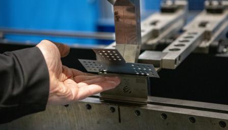 Metallintyöstöprosessi: henkilön käsi pitelee rei'itettyä metallilevyä taivutuskoneessa teollisuusympäristössä.