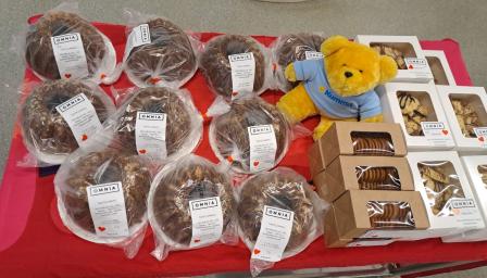 Leivonnaisten myyntipöytä, jossa on Omnian valmistamia tuotteita, kuten taatelikakkuja, pikkuleipiä ja muita herkkuja. Pöydän koristeena on keltainen Kummi-nalle, ja tuotteet on asetettu punaiselle pöytäliinalle.