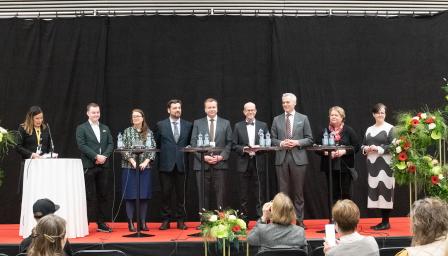 Joukko siististi pukeutuneita ihmisiä seisoo lavalla. Heillä on edessään pyöreät pystypöydät. Lavan edessä näkyy ihmisiä istumassa tuoleilla. Vasemmalla henkilö juontaa tilaisuutta.