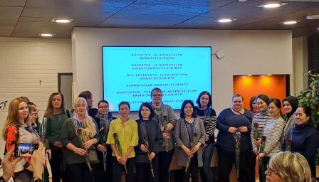 Ryhmä ihmisiä seisoo lavalla rivissä juhlavasti kukkia käsissään. Taustalla valkokankaalle heijastettuna luettelo tutkintojen nimistä, kuten "kasvatus- ja ohjausalan ammattitutkinto" ja "mielenterveys- ja päihdetyön ammattitutkinto". Etualalla yleisöä ottamassa kuvia ja seuraamassa tilaisuutta.