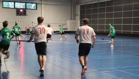 Salibandyottelu meneillään sisäliikuntahallissa. Pelaajat valkoisissa ja vihreissä pelipaidoissa juoksevat kohti maalia, jossa maalivahti odottaa. Etualalla kaksi valkopaitaista pelaajaa, joiden selässä numerot 20 ja 17. Pelikenttä on sininen ja taustalla näkyy tulostaulu.