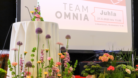 Puhujapönttö ja näyttämö koristeltuna värikkäillä kevätkukkasilla Team Omnian juhlatilaisuudessa. Taustalla suuri näyttö, jossa lukee ”Team Omnia Juhla 27.5.2025 klo 13.00, Liikuntasali, Kirkkokatu 16 B”.