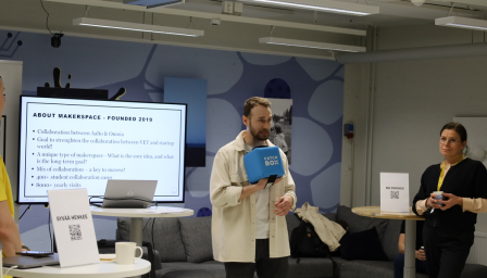 Mies puhuu yleisölle pitäen kädessään sinistä Catchbox-mikrofonikuutiota esitystilanteessa. Taustalla olevassa näytössä näkyy otsikko "ABOUT MAKERSPACE – FOUNDED 2019" ja tietoa Aalto-yliopiston ja Omnian yhteistyöstä. Vasemmalla henkilö kuuntelee esitystä pöydän vieressä, jolla on kyltti ”SYVÄÄ HENKEE” ja QR-koodi. Oikealla pöytä, jossa kyltti ”RD PHYSICS”, sekä kaksi kuuntelijaa. Taustalla näkyy sohva ja seinätaidetta.