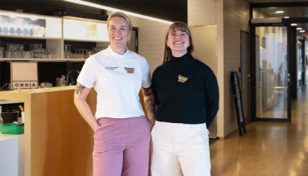 Linnea Vihonen ja Laura Styyra seisovat baaritiskin edessä. Taustalla näkyy Omnian logo ja opetusravintolan tilat. Vihonen ja Styyra katsovat kameraan ja hymyilevät. Vihosella on päällään kokkivaatteet. Molemmilla on rintaneula, jossa lukee Dress to Skill. 