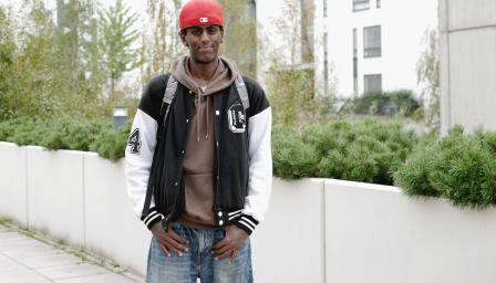 Yacqub Mohamed seisoo pihalla reppu selässä ja hymyilee. Taustalla näkyy kerrostaloja ja vihreää kasvillisuutta.