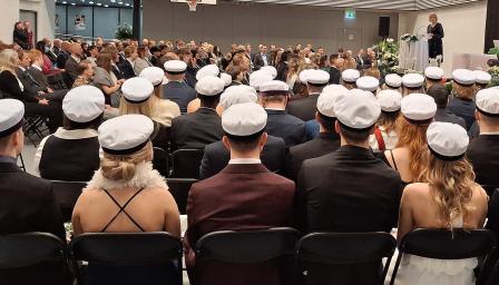 Joukko ylioppilaita istuu liikuntasalissa ylioppilaslakit päässä selkä kameraan päin. Taustalla näkyy puheenpitäjä ja muita ihmisiä.