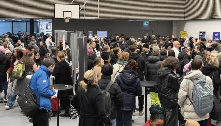 Sisätiloissa järjestetty tapahtuma, jossa suuri joukko ihmisiä seisoo tiiviisti esittelypisteiden ja roll-upien ympärillä; osallistujilla on talvivaatteet ja reppuja, ja tila muistuttaa liikuntasalia.