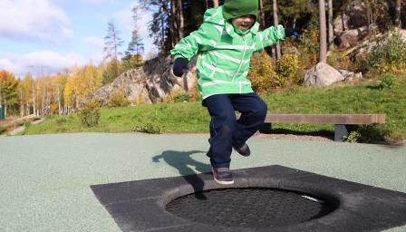 Vihreään takkin ja lakkiin pukeutunut lapsi hyppii trampoliinilla puistossa. Taustalla kallioista metsää.