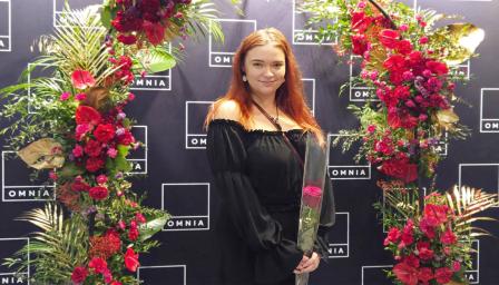 Ronja Oksanen seisoo kukkakaaren alla ruusu kädessään ja hymyilee.