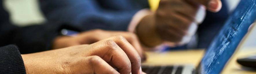 Opiskelija käyttää tietokonetta ja taustalla ohjaaja osoittaa ruudulta asiaa.