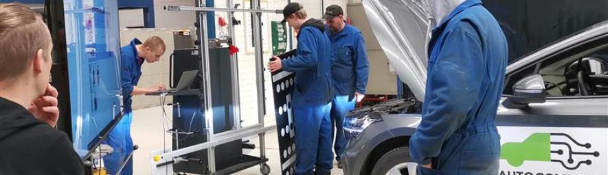Autoalan opiskelijoita työhaalareissa tutkimassa auton teknisiä järjestelmiä korjaamoympäristössä. Etualalla näkyvässä autossa on "AUTOCOVE 2.0" -logo ja konepelti auki huoltoa varten.