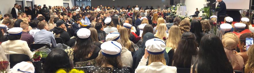 Suuri joukko ihmisiä istuu jumppasalissa juhlavaatteissa selkä kohti kameraa. Osalla on päässään valkoinen ammattilaislakki.