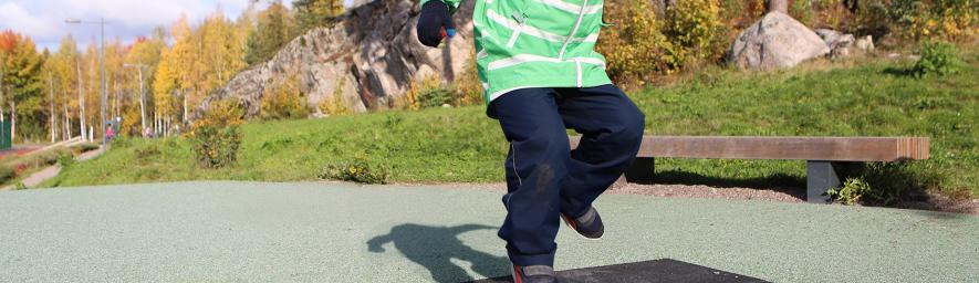 Vihreään takkin ja lakkiin pukeutunut lapsi hyppii trampoliinilla puistossa. Taustalla kallioista metsää.