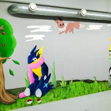 Seinämaalaus, jossa näkyy useita Pokémon-hahmoja kuten Greninja, Eevee, Pikachu ja Bulbasaur, sekä sarjakuvamainen puu ja Poképalloja vihreässä ruohomaisemassa.