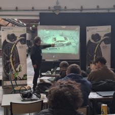 Auto CoVE 2.0 -hankkeen työpajassa opettaja esittelee auton kalibrointia valkokankaalla. Oppilaat seuraavat esitystä pöytien ääressä, ja taustalla näkyy hankkeen roll-up julisteineen.