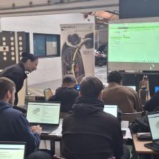 Auto CoVE 2.0 -hankkeen työpajassa opiskelijat työskentelevät kannettavien tietokoneiden äärellä. Opettaja neuvoo opiskelijoita, ja valkokankaalla näkyy Electude-oppimisalustan näkymä.