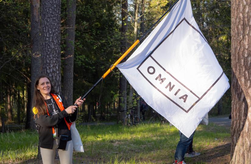 Nina Kauppinen seisoo kaupunkimetsässä ja pitää kädessään Omnian lippua.