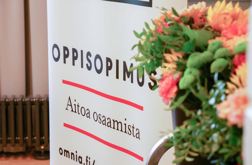 Taustalla roll-up, jossa teksti Oppisopimus, aitoa osaamista. Etualalla kukkia.