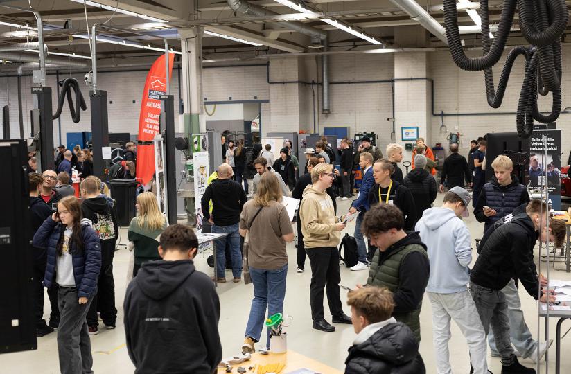 Suuri määrä nuoria ihmisiä tutustumassa autoalan edustajien ständeihin. Ympäristönä on autohalli.