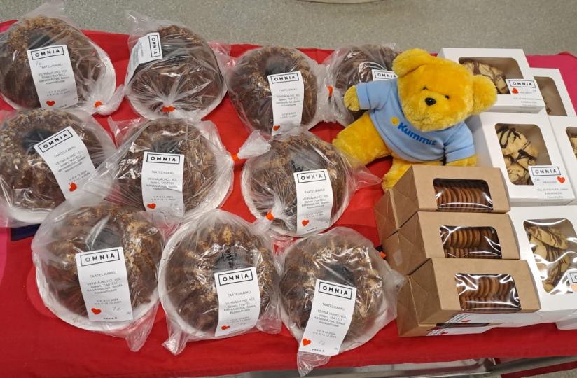 Leivonnaisten myyntipöytä, jossa on Omnian valmistamia tuotteita, kuten taatelikakkuja, pikkuleipiä ja muita herkkuja. Pöydän koristeena on keltainen Kummi-nalle, ja tuotteet on asetettu punaiselle pöytäliinalle.