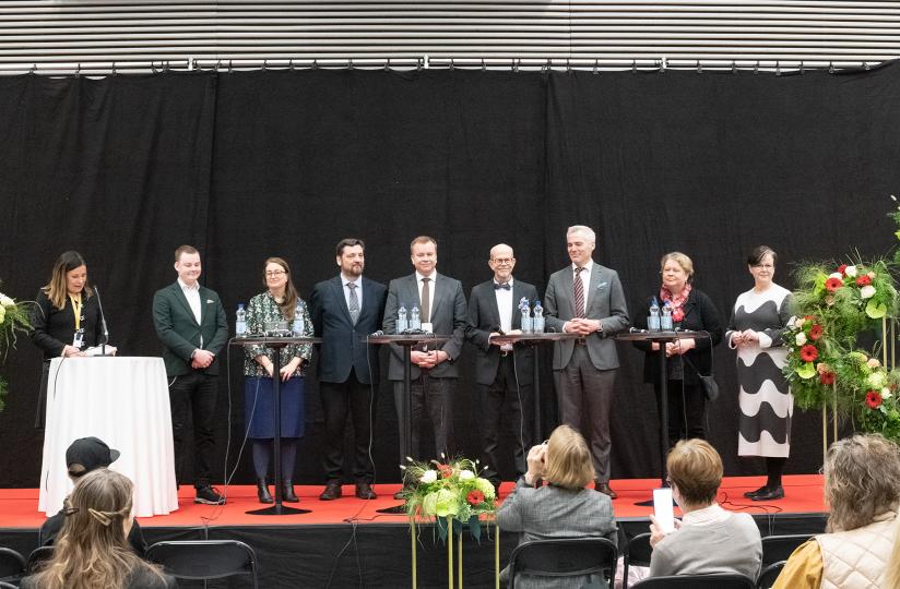 Joukko siististi pukeutuneita ihmisiä seisoo lavalla. Heillä on edessään pyöreät pystypöydät. Lavan edessä näkyy ihmisiä istumassa tuoleilla. Vasemmalla henkilö juontaa tilaisuutta.