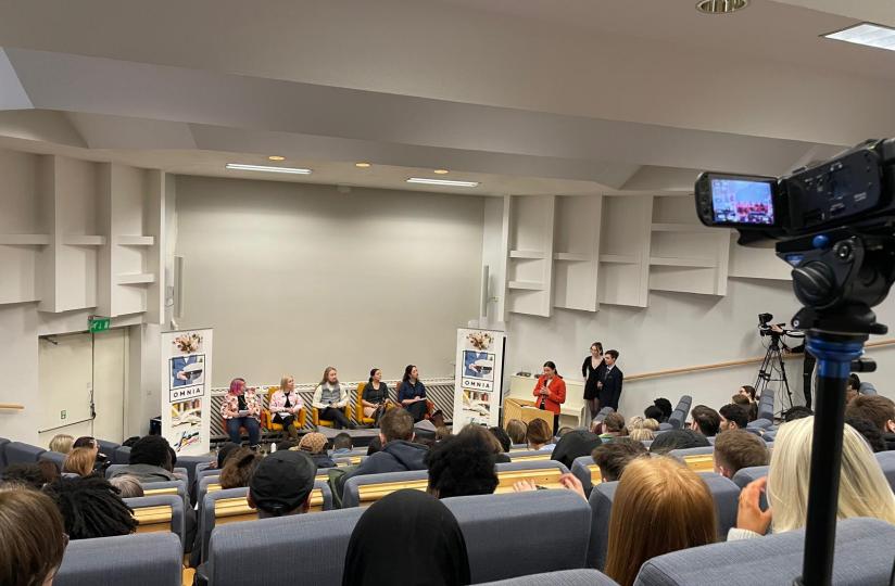 Yleisö seuraa vaalipaneelia auditorioympäristössä. Panelistit istuvat lavalla Omnian mainosjulisteiden edessä, ja esiintyjä seisoo puhujanpöntössä. Tilaisuutta taltioidaan videokameralla, joka näkyy kuvan oikeassa laidassa.