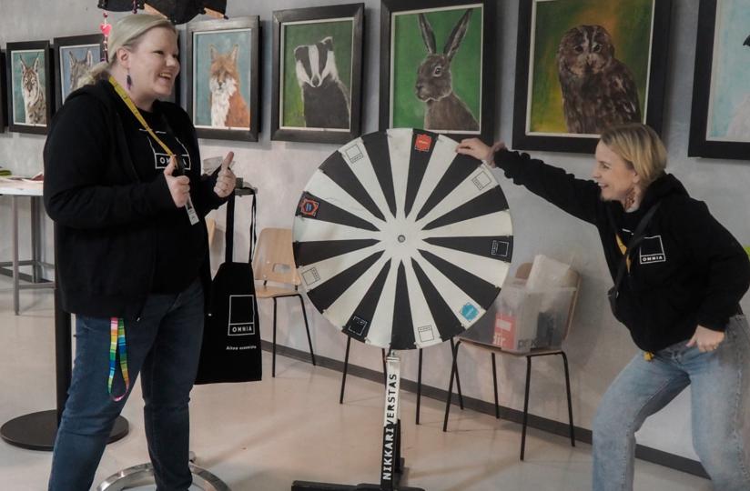 Kaksi henkilöä Omnia-huppareissa seisoo sisätiloissa onnenpyörän äärellä iloisina. Toinen pitää kiinni pyörästä ja toinen näyttää peukkua. Taustalla on eläinaiheisia maalauksia seinällä sekä tuoleja ja pöytiä, joilla on materiaaleja. Pyörässä lukee "Nikkariverstas".