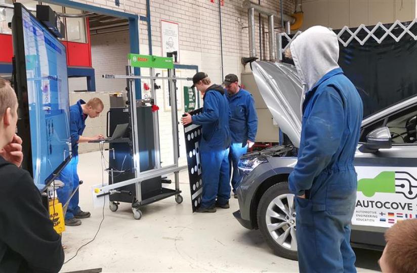 Autoalan opiskelijoita työhaalareissa tutkimassa auton teknisiä järjestelmiä korjaamoympäristössä. Etualalla näkyvässä autossa on "AUTOCOVE 2.0" -logo ja konepelti auki huoltoa varten.