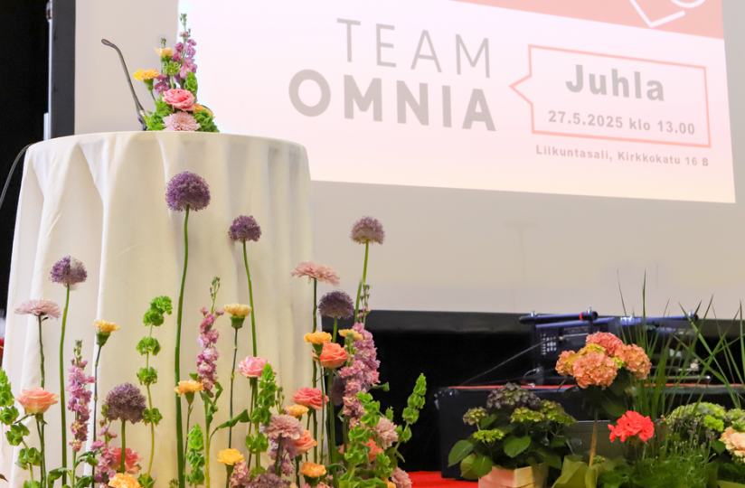 Puhujapönttö ja näyttämö koristeltuna värikkäillä kevätkukkasilla Team Omnian juhlatilaisuudessa. Taustalla suuri näyttö, jossa lukee ”Team Omnia Juhla 27.5.2025 klo 13.00, Liikuntasali, Kirkkokatu 16 B”.