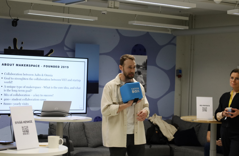 Mies puhuu yleisölle pitäen kädessään sinistä Catchbox-mikrofonikuutiota esitystilanteessa. Taustalla olevassa näytössä näkyy otsikko "ABOUT MAKERSPACE – FOUNDED 2019" ja tietoa Aalto-yliopiston ja Omnian yhteistyöstä. Vasemmalla henkilö kuuntelee esitystä pöydän vieressä, jolla on kyltti ”SYVÄÄ HENKEE” ja QR-koodi. Oikealla pöytä, jossa kyltti ”RD PHYSICS”, sekä kaksi kuuntelijaa. Taustalla näkyy sohva ja seinätaidetta.