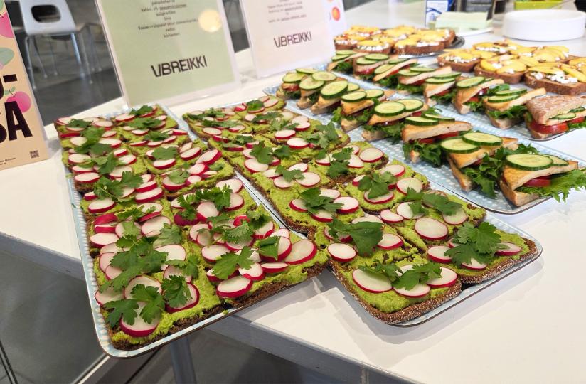 Tarjoilupöydällä on riveittäin koristeltuja voileipiä, joista osa on päällystetty vihreällä levitteellä, retiisiviipaleilla ja korianterilla. Toisilla leivillä on salaattia, tomaattia, paistettua tofua tai juustoa ja kurkkua. Taustalla näkyy kylttejä, joissa on VBREIKKI-logo ja leipien täytteiden tiedot.