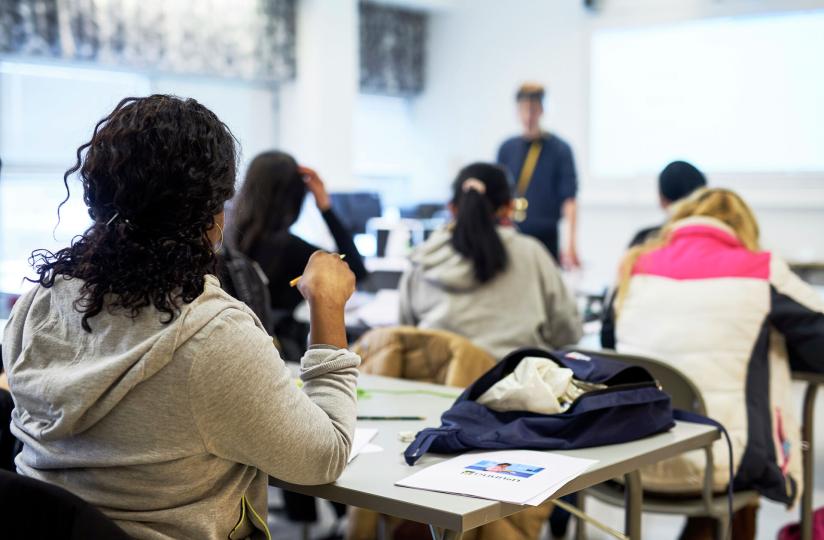 Ryhmä aikuisopiskelijoita seuraa opetusta luokkahuoneessa, jossa opettaja seisoo valkotaulun edessä. Etualalla näkyy opiskelija harmaassa hupparissa ja pöydällä Omnian esite.