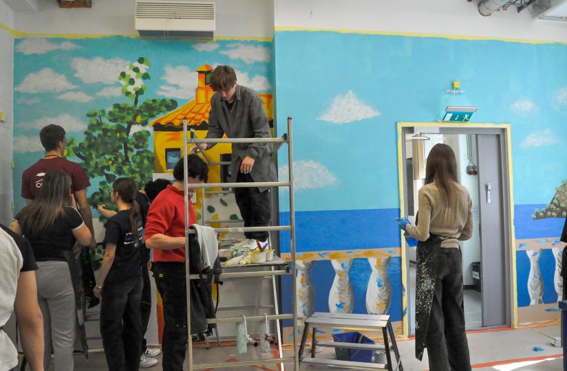 Useat nuoret maalaavat yhdessä värikästä seinämaalausta sisätiloissa. Vasemmalla ryhmä työstää vihreitä puita ja keltaista taloa sinitaivasta vasten, kun taas oikealla puolella näkyy sininen merimaisema ja pylväsaiheinen parveke. Keskellä nuori seisoo maalaustelineellä, ja toinen henkilö katsoo seinää maalausessu päällään. Tunnelma on keskittynyt ja luova.