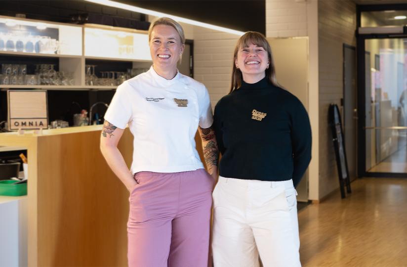 Linnea Vihonen ja Laura Styyra seisovat baaritiskin edessä. Taustalla näkyy Omnian logo ja opetusravintolan tilat. Vihonen ja Styyra katsovat kameraan ja hymyilevät. Vihosella on päällään kokkivaatteet. Molemmilla on rintaneula, jossa lukee Dress to Skill.