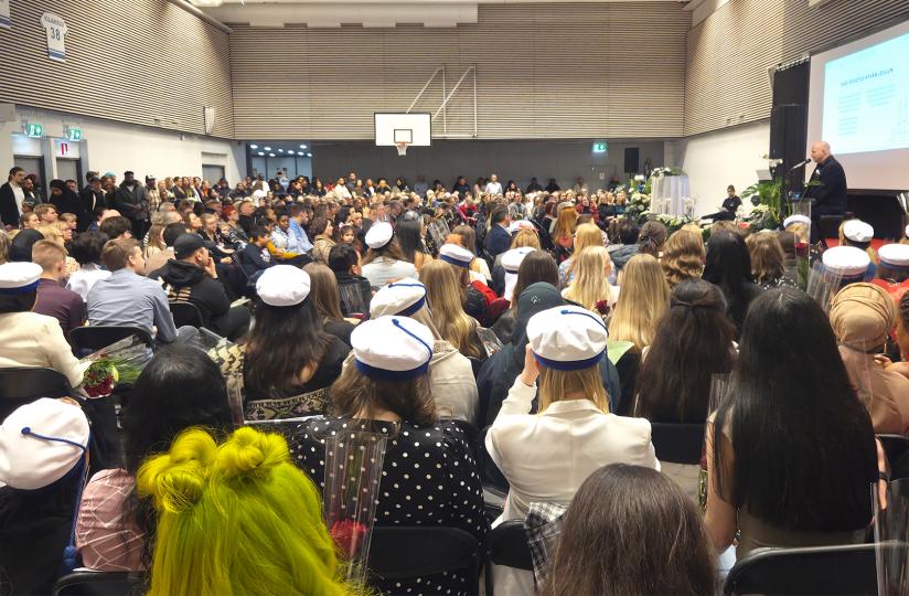 Suuri joukko ihmisiä istuu jumppasalissa juhlavaatteissa selkä kohti kameraa. Osalla on päässään valkoinen ammattilaislakki.