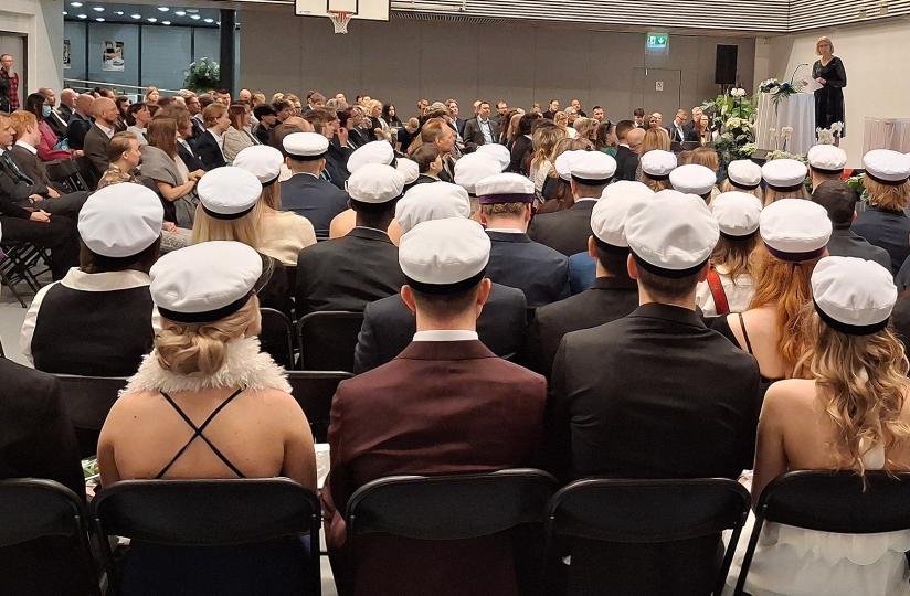 Joukko ylioppilaita istuu liikuntasalissa ylioppilaslakit päässä selkä kameraan päin. Taustalla näkyy puheenpitäjä ja muita ihmisiä.