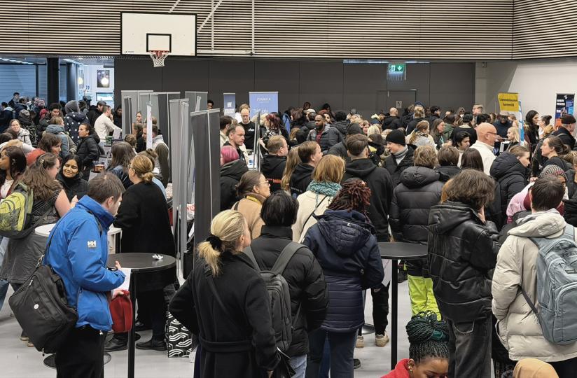 Sisätiloissa järjestetty tapahtuma, jossa suuri joukko ihmisiä seisoo tiiviisti esittelypisteiden ja roll-upien ympärillä; osallistujilla on talvivaatteet ja reppuja, ja tila muistuttaa liikuntasalia.