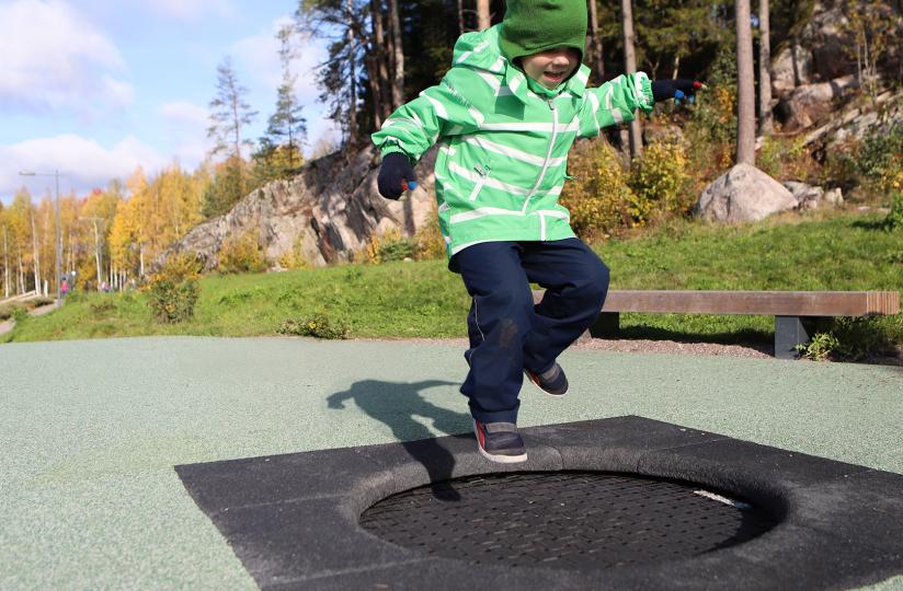 Vihreään takkin ja lakkiin pukeutunut lapsi hyppii trampoliinilla puistossa. Taustalla kallioista metsää.