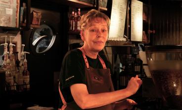 Päivi Järvinen seisoo kahvikoneen takana ja katsoo kameraan.
