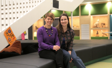 Paula Havu ja Siiri Autio istuvat koulun aulassa sohvalla ja katsovat kameraan.