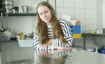 Hanne Jauhiainen nojaa keittiön työtasoon ja katsoo kameraan. Taustalla näkyy keittiön työvälineitä.