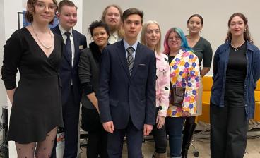 Ryhmä nuoria seisoo yhdessä virallisessa tilaisuudessa, osa pukeutuneena jakkupukuun ja osa värikkäisiin asuihin. Taustalla näkyy Omnia-brändätty esittelyjuliste. Kuva on otettu sisätiloissa vaalipaneelin yhteydessä.