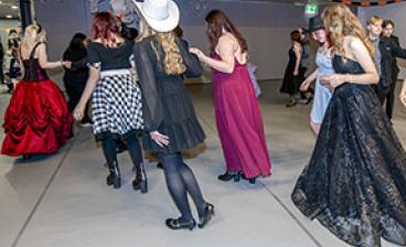 Opiskelijoita tanssimassa juhlasalissa juhlapuvuissa, osa pukeutunut myös teema-asuihin kuten valkoiseen cowboy-hattuun. Tunnelma on iloinen ja rennon juhlava.