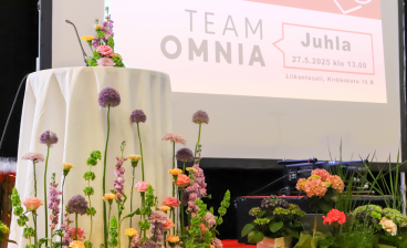 Puhujapönttö ja näyttämö koristeltuna värikkäillä kevätkukkasilla Team Omnian juhlatilaisuudessa. Taustalla suuri näyttö, jossa lukee ”Team Omnia Juhla 27.5.2025 klo 13.00, Liikuntasali, Kirkkokatu 16 B”.