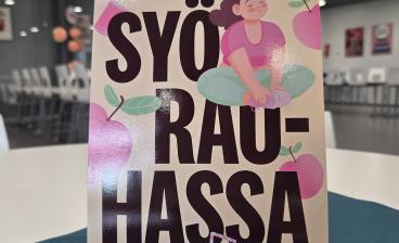 Pöydällä sinisen tabletin päällä on pystyssä oleva kampanjajuliste, jossa lukee isolla “Syö rauhassa”. Julisteessa on myös piirros meditoivasta hahmosta sekä vaaleanpunaisia hedelmäkuvituksia. Taustalla näkyy ruokasali, jossa on tyhjiä valkoisia tuoleja ja pöytiä.