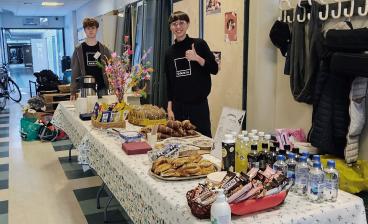 Kahvilapöytä kuvattuna sivulta. Pöydän ääressä seisoo kaksi opiskelijaa, toinen antaa peukun merkiksi. Tarjoilut sisältävät pullia, suklaata ja juomia.